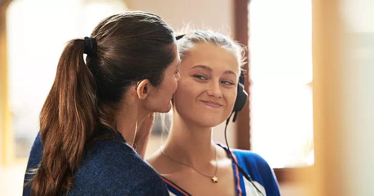 Due donne in un ambiente luminoso, una con capelli castani raccolti e una collana a stella, mentre interagiscono. L’immagine non mostra chiaramente apparecchi acustici né loghi specifici.
