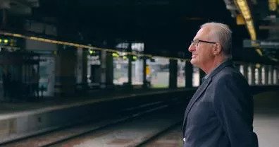 Uomo anziano in piedi sulla banchina di una stazione ferroviaria.