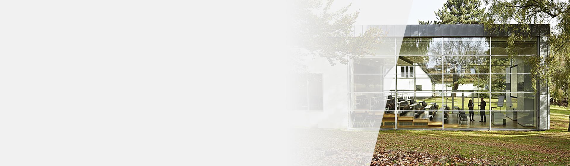 Edificio moderno con facciata in vetro, riflettente un giardino verde circostante; all’interno si vedono due persone conversare in un’aula luminosa arredata con sedie e dispositivi audiovisivi.