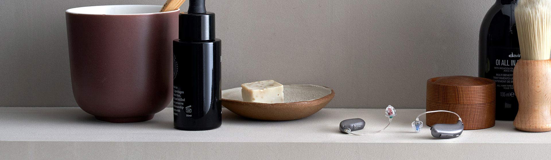 Due apparecchi acustici moderni sono posati su una superficie liscia accanto a oggetti da bagno: una tazza in ceramica, una bottiglia nera, sapone, un pennello e contenitori in legno.
