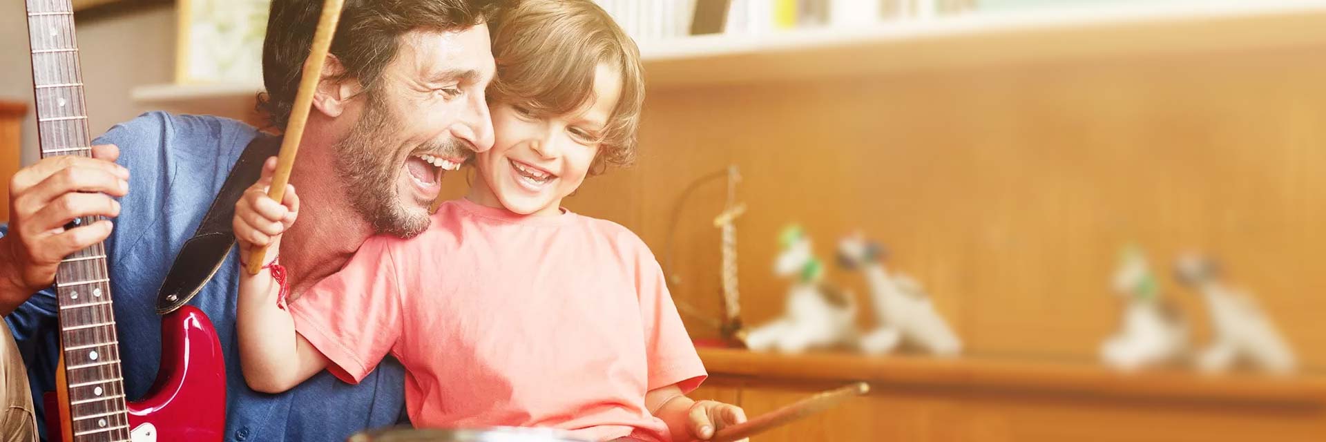 Una chitarra elettrica rossa è suonata da un adulto accanto a un bambino che usa bacchette per batteria. Lo sfondo mostra scaffali in legno con decorazioni e oggetti.