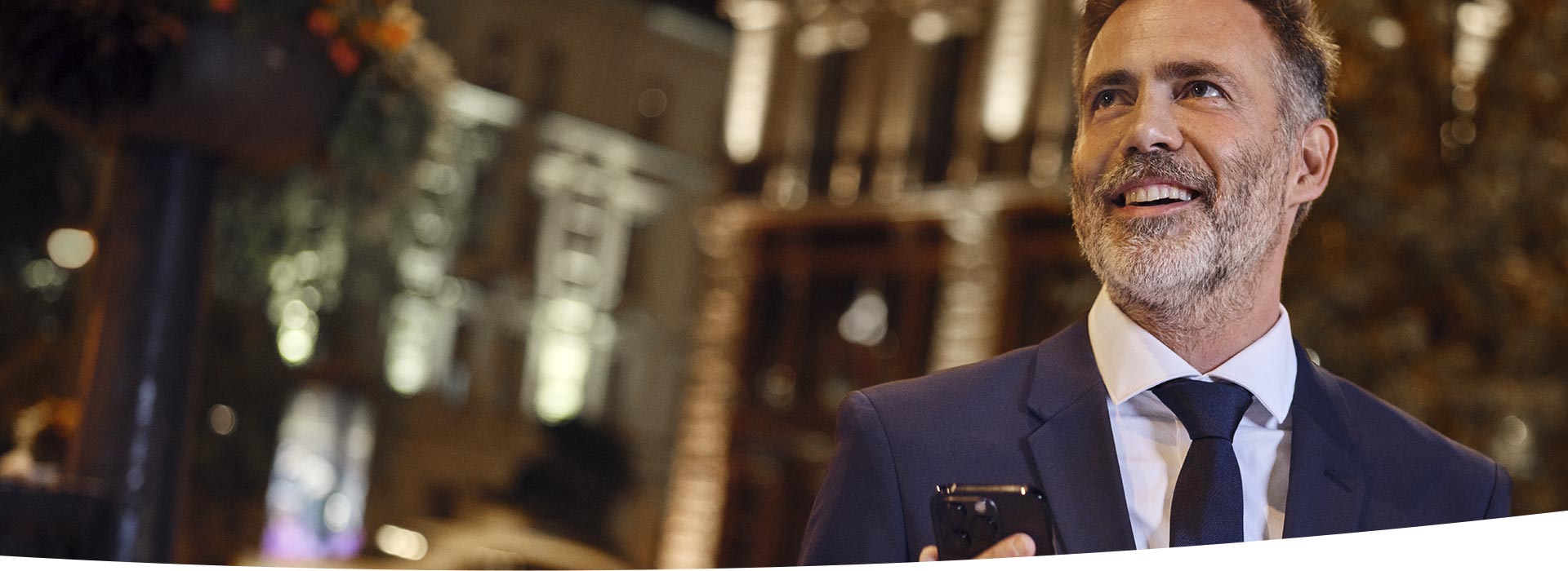 Un uomo in abito elegante tiene uno smartphone in mano, in un contesto urbano notturno con edifici illuminati e luci sfocate sullo sfondo. Nessun testo visibile.