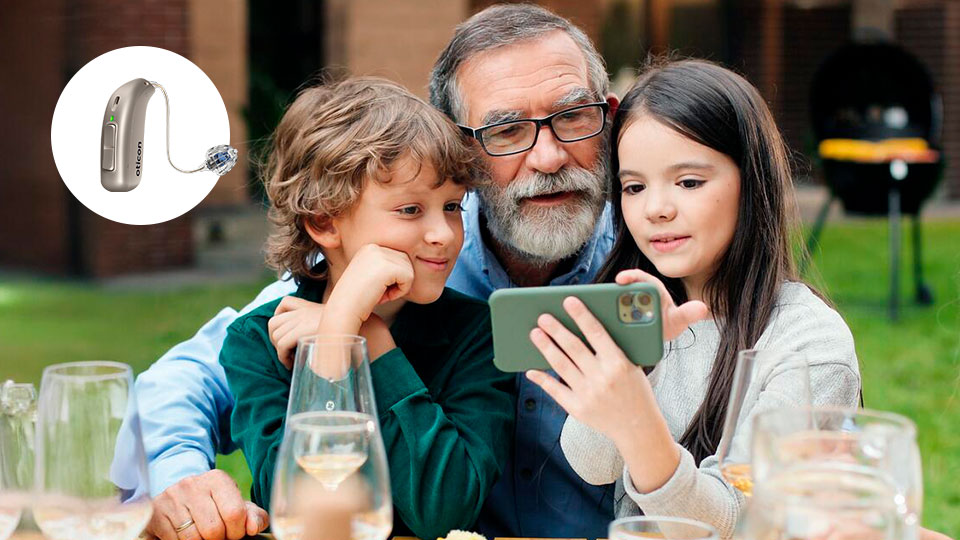 Un apparecchio acustico Oticon è mostrato a sinistra. Sullo sfondo, una famiglia osserva uno smartphone in un giardino all’aperto con bicchieri e barbecue visibili.