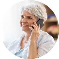 Una donna anziana dai capelli bianchi parla al telefono, seduta in un ambiente domestico luminoso; sullo sfondo appaiono sfocati mobili in legno e decorazioni floreali. Nessun marchio o testo visibile.