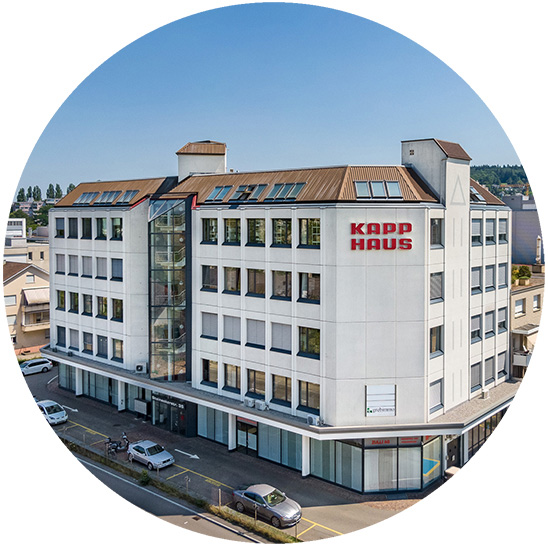 L’edificio principale è un complesso commerciale chiamato ’KAPP HAUS’ con cinque piani, finestre ampie e un’entrata centrale in vetro; situato in una zona urbana con auto parcheggiate.