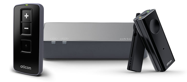 Tre dispositivi Oticon: un telecomando nero con pulsanti '+' e '-', una base grigia con logo Oticon e un microfono nero portatile, progettati per supportare apparecchi acustici.