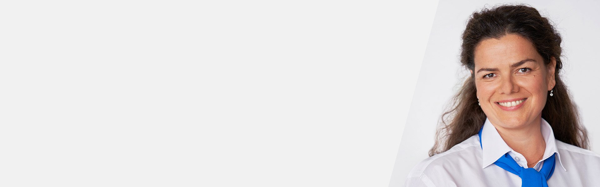 Una donna con capelli ricci castani indossa una camicia bianca con cravatta blu. Sfondo bianco minimalista, senza testo visibile né apparecchi acustici o marchi identificabili.