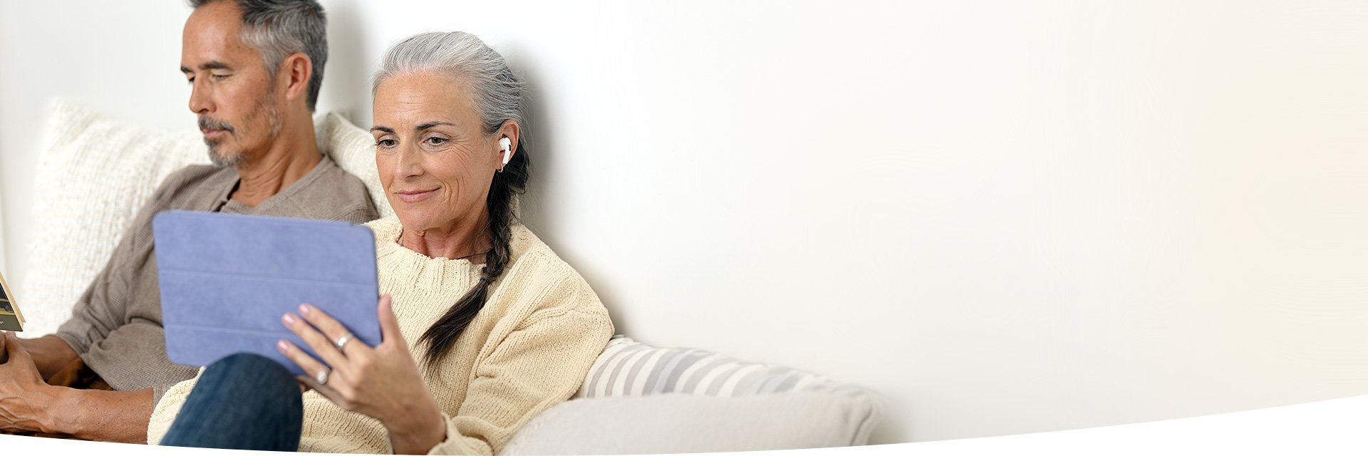Donna con capelli grigi utilizza auricolari mentre guarda un tablet seduta accanto a un uomo su un divano.