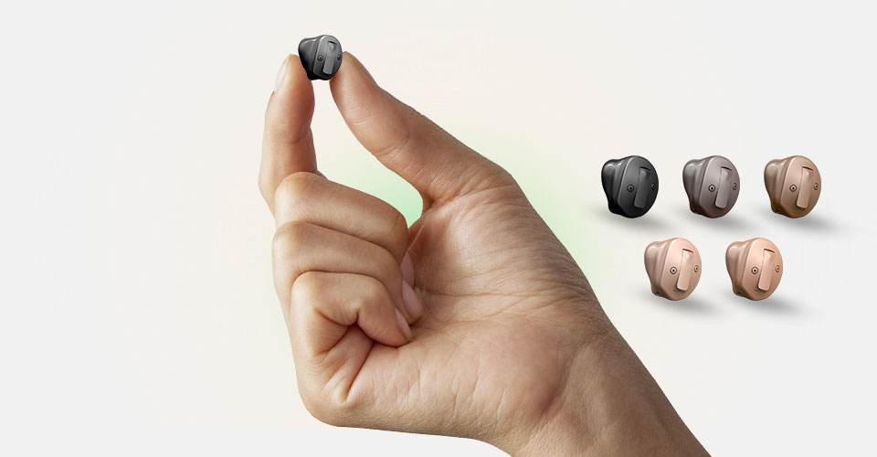 Una mano tiene delicatamente un apparecchio acustico discreto e interno, circondato da diverse varianti di colore disposte su sfondo chiaro, suggerendo opzioni di personalizzazione per l’udito.