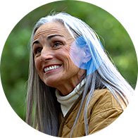 Una persona con capelli lunghi grigi indossa una giacca marrone e si trova all’aperto con uno sfondo verde. Sul lato dell’orecchio è visibile un apparecchio acustico azzurro.