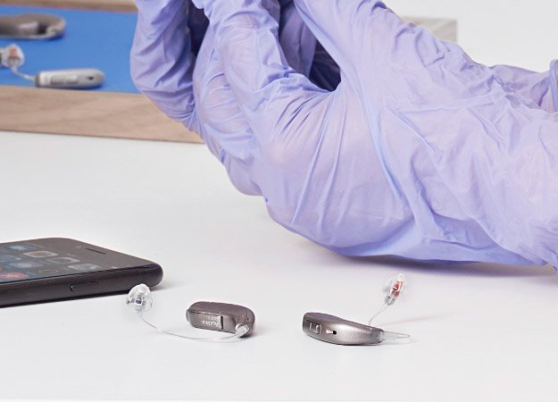 Dispositivi acustici moderni su tavolo con smartphone, assistenza professionale suggerita da mani guantate viola.
