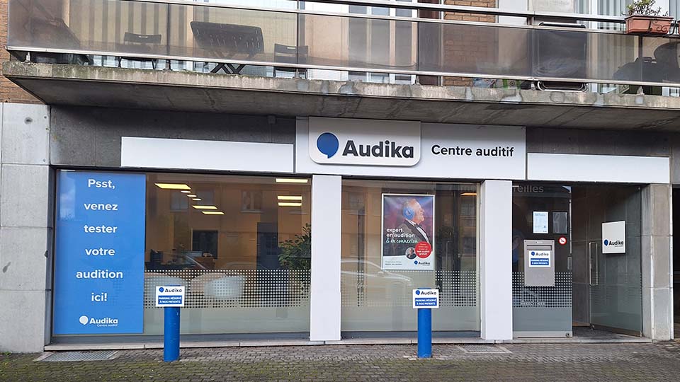 De gevel van een Audika-hoorcentrum toont branding met teksten: ’Psst, venez tester votre audition ici!’ en ’Centre auditif.’ Binnenin zijn verlichte ruimtes zichtbaar, met een poster over hooroplossingen.