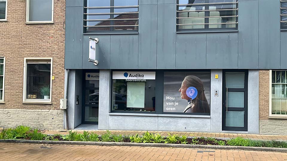 De voorgevel van een Audika hoorcentrum toont een glazen ingang, een raam met het logo en opschrift ’Audika hoorcentrum,’ en een poster met de tekst ’Hou van je oren.’