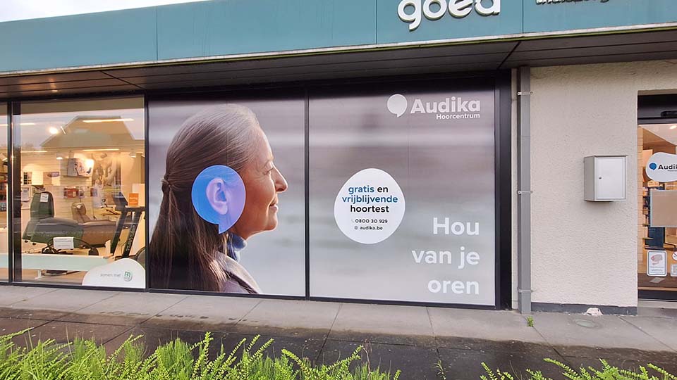 Winkelgevel van Audika Hoorcentrum met reclametekst: ’gratis en vrijblijvende hoortest 0800 30 929 audika.be’ en ’Hou van je oren.’ Binnenin zichtbare audiologische apparatuur en displays.