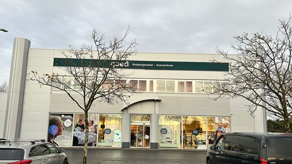Een zorgwinkel met de naam 'Pedo' biedt thuiszorgproducten en een hoorcentrum. Grote ramen tonen binnenactiviteiten, en buiten staan auto's geparkeerd. Links staat een poster met de tekst: 'Audika - Hou van je oren.' Boomtakken in wintersfeer omlijsten het gebouw.