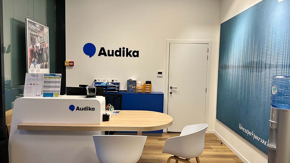 Een moderne hoorzorgbalie met het Audika-logo, folders over hoorzorg, een wachtruimte met witte stoelen en een blauwe muurtekst: 'Votre expert pour une...' naast een waterdispenser.