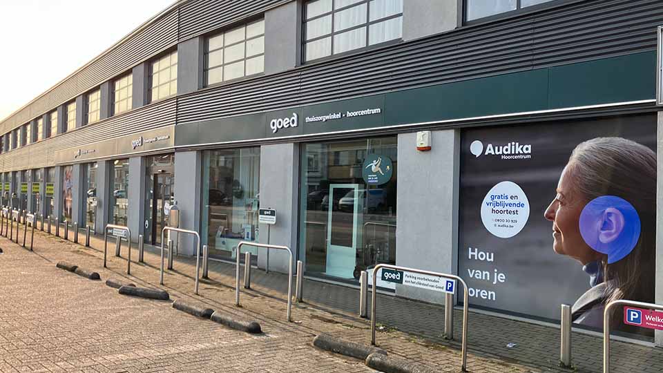Gevel van een hoorzorgwinkel met het logo ’Audika hoorcentrum’ en de tekst: ’Gratis en vrijblijvende hoortest. 0800 50 30’; slogan: ’Hou van je oren.’ Rechts parkeerplekcontext.