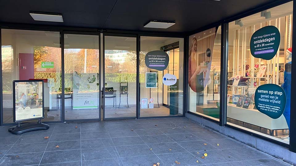 De Audika-hoorwinkel toont glazen ramen met teksten zoals: 'Ontdekdagen op 25 & 26 november' en 'samen op stap, geniet van je mobiele vrijheid,' omlijst door een moderne kliniek-setting.