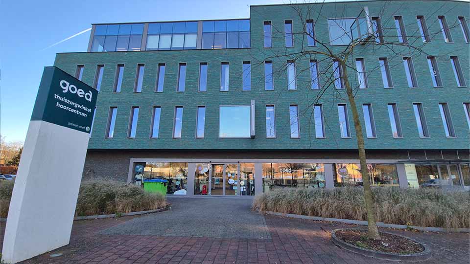 Het gebouw heeft een moderne gevel met smalle ramen en groene bakstenen. Op de voorgrond staat een bord met tekst: 'goed thuiszorgwinkel hoorcentrum samen wel'. De ingang leidt naar een hoorcentrum voor gehoorzorg.