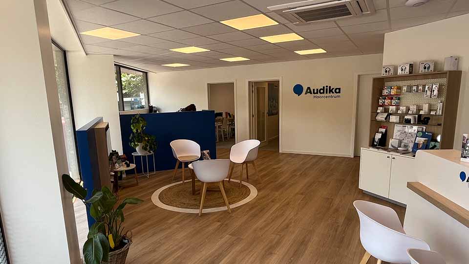 Een moderne wachtruimte met houten vloer, witte stoelen en een ronde mat. Aan de muur staat 'Audika Hoorcentrum'. Rechts een kast met hoorapparaatproducten en folders.