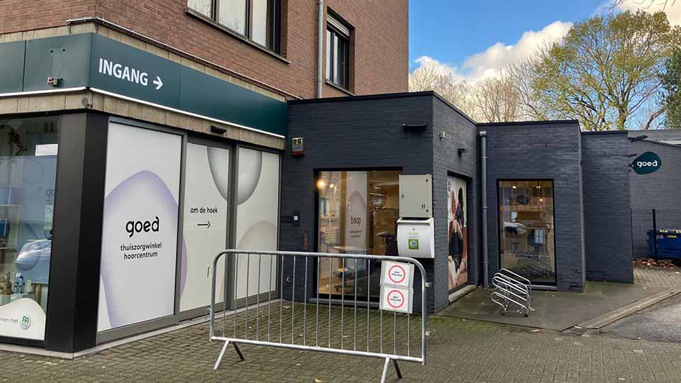 Een hoorzorgcentrum met de tekst 'goed thuiszorgwinkel hoorcentrum' op de ramen. De ingang, aangegeven met een bord 'INGANG →', bevindt zich in een nette, modern ogende straatomgeving met bomen op de achtergrond.