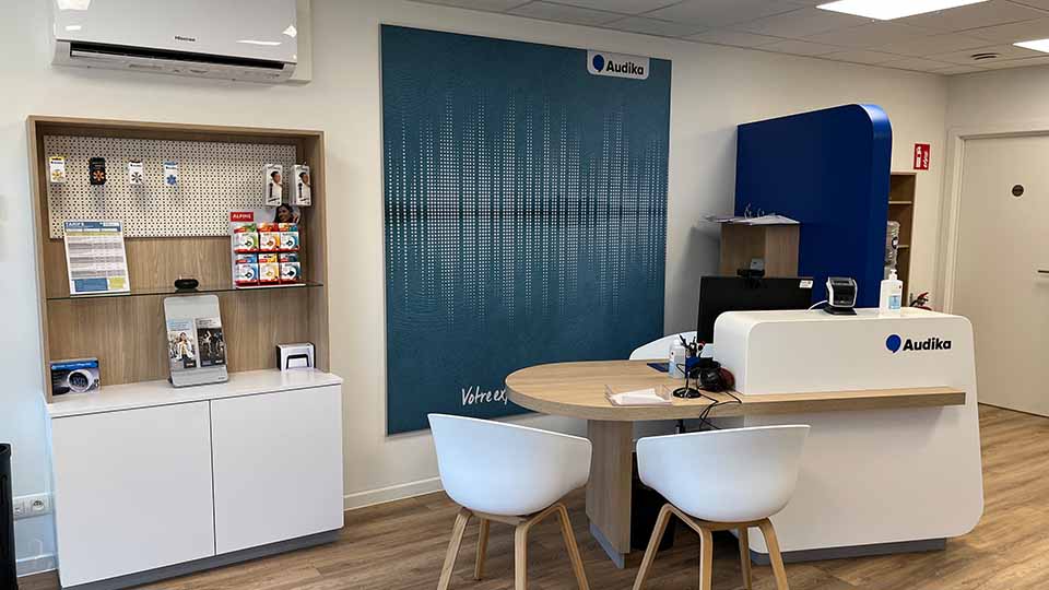 Een moderne hoorwinkel van Audika met een consultatiedesk, stoelen en promotiemateriaal. Links vitrine met hoortoestelaccessoires, rechts een blauwe wand met het Audika-logo.