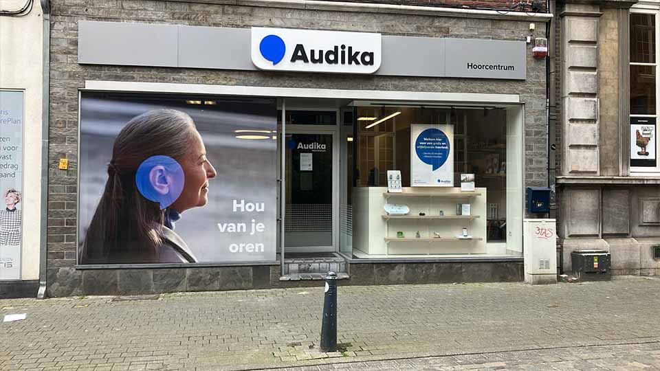 De voorgevel van het Audika Hoorcentrum toont links een grote etalage met een poster van een persoon met een blauw oor en de tekst 'Hou van je oren.' Rechts zijn een deur met het logo 'Audika' en een etalage met hoortoestellen en een informatieve tekst zichtbaar. Gelegen aan een straat in een gebouw met stenen gevel.