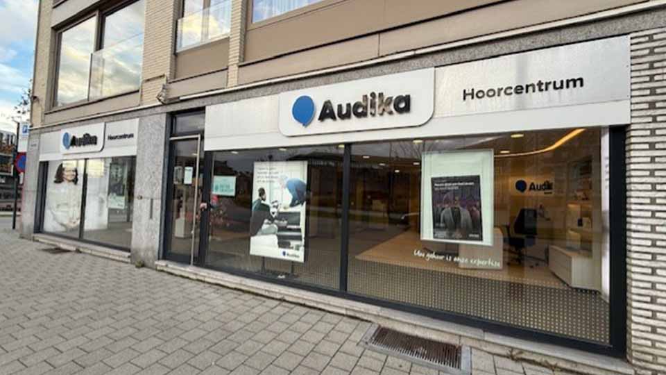 Audika hoorcentrum met glazen etalage toont posters over gehoorzorg, waaronder een audioloog die een gehoortest uitvoert. Exterieur met trottoir, grijze stenen gevel en straatzicht.