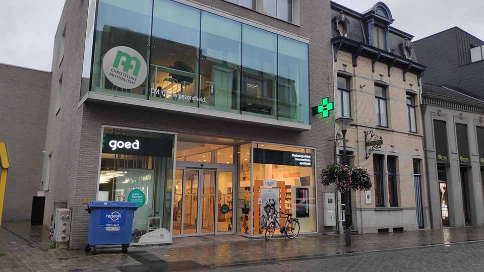 Een modern gebouwencomplex met een glazen gevel toont winkels, waaronder 'goed' en een apotheek met een groen kruis. Context: natte straat, fiets en bloembakken. Tekst: 'Christelijke Mutualiteit' en 'www.gezondheid.be'.