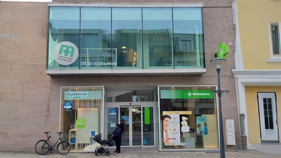 Een modern gebouwencomplex met een glazen gevel toont winkels, waaronder 'goed' en een apotheek met een groen kruis. Context: natte straat, fiets en bloembakken. 