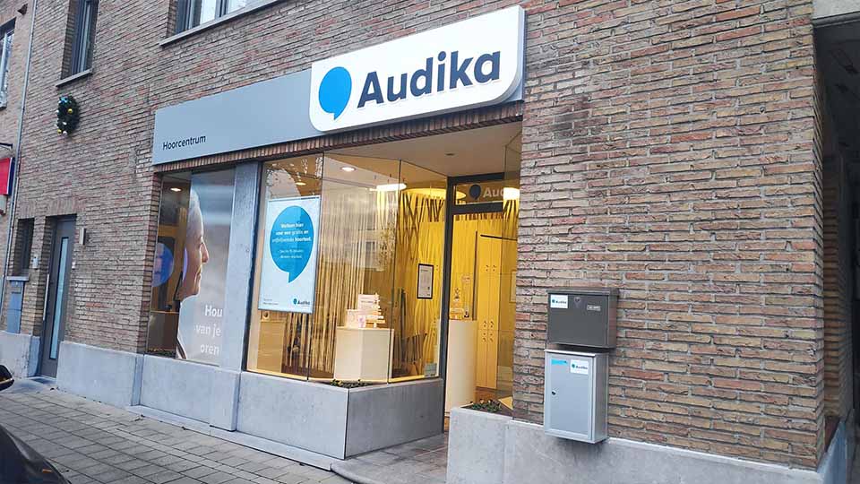 De voorgevel van een Audika hoorcentrum met glazen ramen toont een uitnodigende ruimte. Boven de ingang hangt een wit bord met het blauwe logo 'Audika'. Links op het raam staat tekst: 'Hou van je oren'.  Context: Gelegen aan een bakstenen gebouw in een rustige straat.