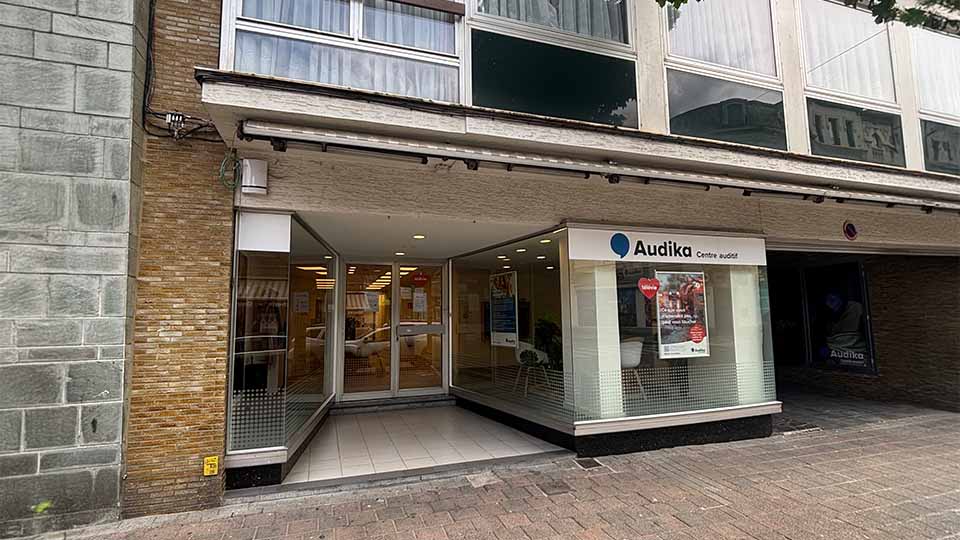 De ingang van een Audika hoorcentrum met glazen schuifdeuren, een helder interieur en een poster over hoorzorgdiensten in de etalage; gevestigd aan een winkelstraat met bakstenen gevel.