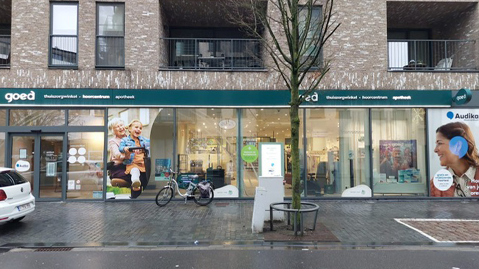 De winkel 'goed' combineert een thuiszorgwinkel, hoorcentrum en apotheek. Achter de glazen gevel zijn medische apparatuur en producten zichtbaar. Links is Audika gepresenteerd, gespecialiseerd in hoorzorg.