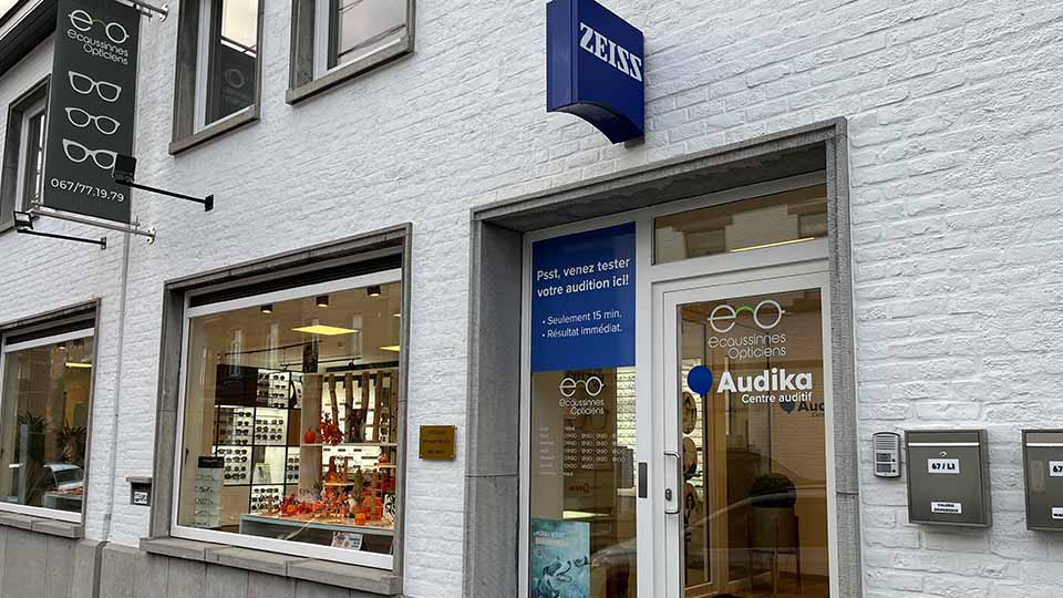 De voorgevel van een Audika hoorcentrum in een wit gebouw, met naastgelegen optiekwinkel ’Ecoussines Opticiens.’ Tekst op de deur: ’Psst, venez tester votre audition ici! - Seulement 15 min. - Résultat immédiat.’