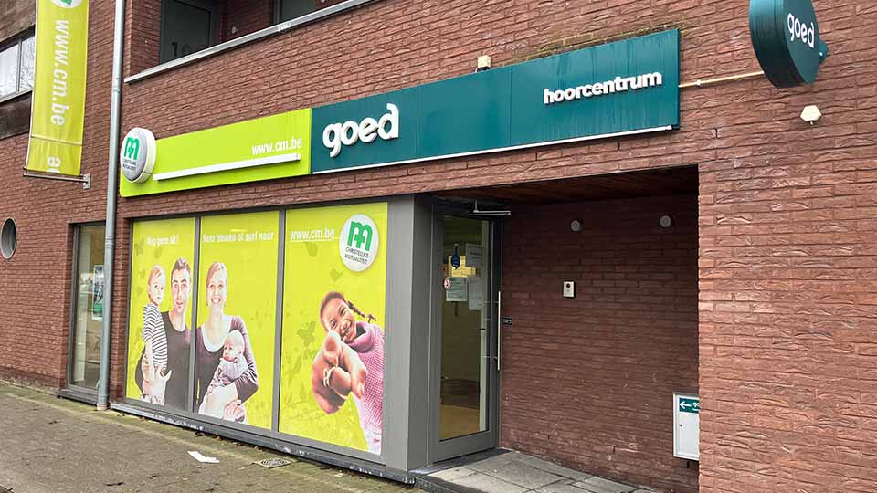 De gevel van een hoorzorgcentrum met de tekst ’goed hoorcentrum’ op een blauwe balk, naast een glazen ingang en etalage met groene reclame van www.cm.be en blurrende gezichten.