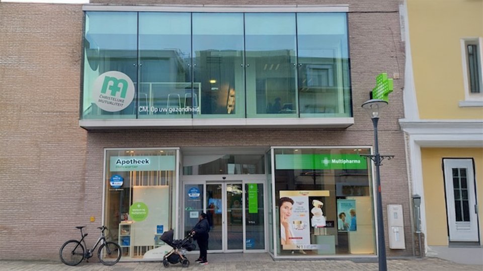 Een modern gebouwencomplex met een glazen gevel toont winkels, waaronder 'goed' en een apotheek met een groen kruis. Context: natte straat, fiets en bloembakken. 