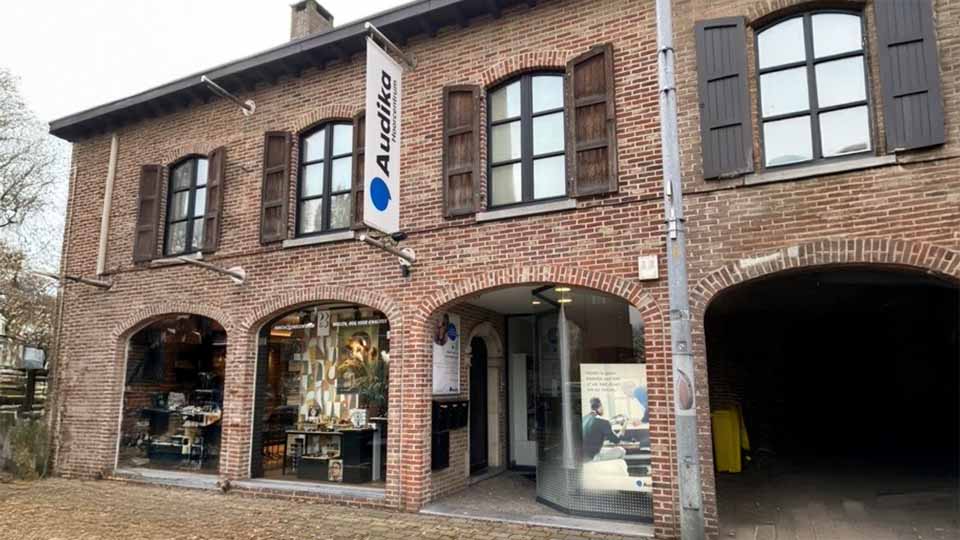 Audika hoortoestellenwinkel in een historisch bakstenen pand met houten luiken. Een bord met ’Audika’ hangt boven de ingang. Binnenin zichtbaar: posters, displays en hoorzorgartikelen.