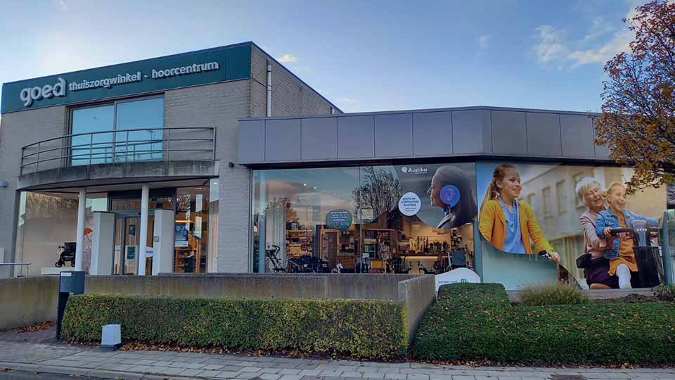 Een hoorzorgwinkel en thuiszorgcentrum gevestigd in een modern gebouw. Bovenaan staat de tekst: ’goed thuiszorgwinkel - hoorcentrum.’ Rechts is het Audika-logo zichtbaar op een glazen gevel met reclameafbeeldingen van mensen met hoortoestellen. De omgeving bevat een stoep, een groene haag en een boom.