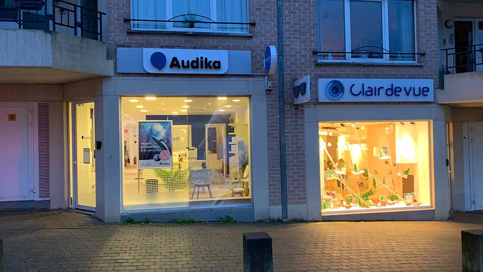 De Audika-gehoorwinkel toont een verlichte etalage met de tekst ’AIMEZ VOS OREILLES.’ Binnen zijn een poster en zitmeubels zichtbaar. Buurpand Clair de Vue biedt brillen in een warme sfeer.