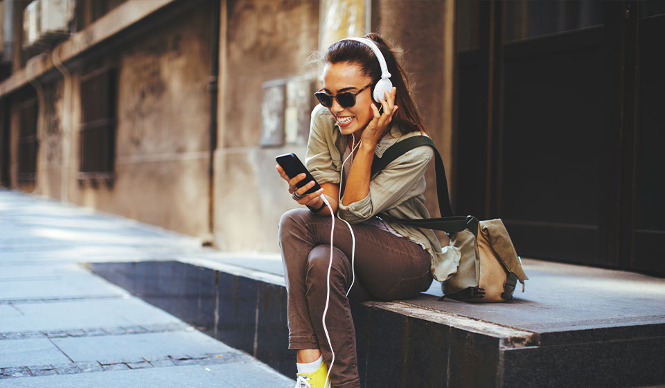 Vrouw met koptelefoon luistert naar muziek op haar telefoon terwijl ze op een stoeprand zit.