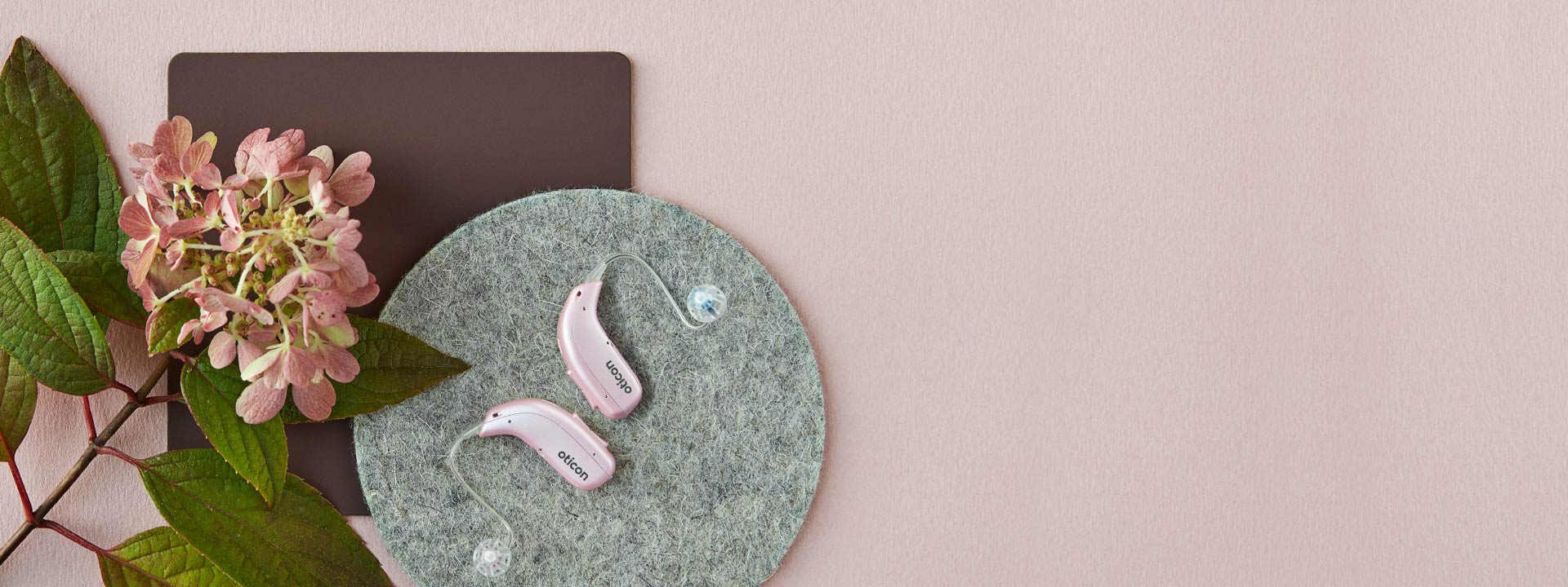Roze Oticon-hoorapparaten op een vilten ondergrond naast een hortensiabloem.