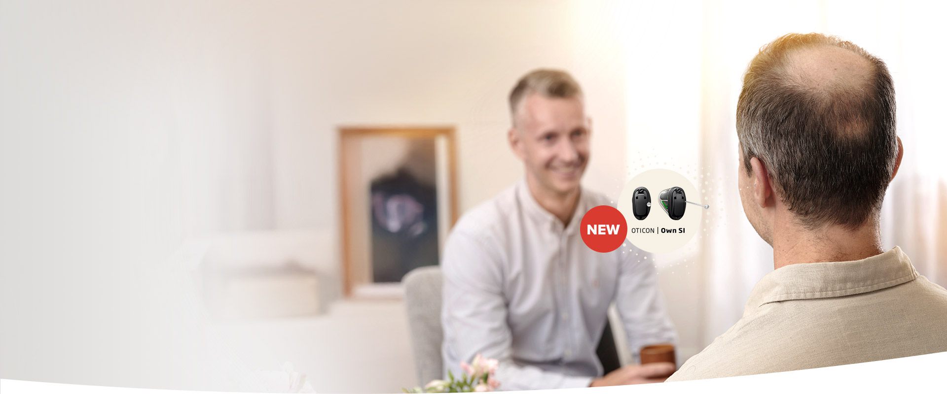 Een man met kort grijs haar zit in een lichte, huiselijke omgeving en spreekt met een audioloog in een wit shirt. Rechts staat tekst: ’NEW Oticon Own SI’ met een afbeelding van Oticon hoortoestellen.
