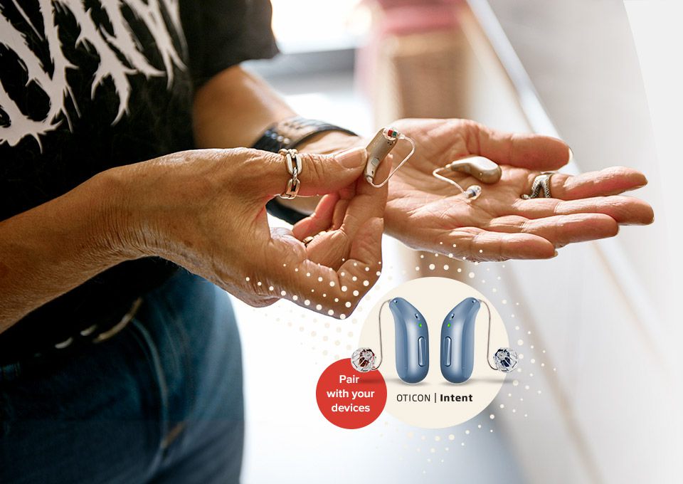 Een persoon houdt twee hoortoestellen in de hand, terwijl Oticon Intent-hoortoestellen worden afgebeeld met de tekst: ’Pair with your devices.’ Context: modern binnenruimte, lichte achtergrond.