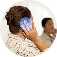 Een vrouw houdt een vergrootglas bij haar oor, met een focus op een gehoorapparaat. Ze staat in een moderne gehoorkliniek; de onscherpe achtergrond toont een andere persoon. Geen zichtbare merknaam.