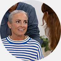 Een oudere persoon met grijs haar draagt een blauw-wit gestreepte trui en zit in gesprek met een audioloog in een professionele, lichte omgeving, mogelijk een gehoorcentrum. Geen zichtbare gehoorapparaten of tekst.