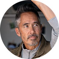 Een man met grijs haar draagt een casual outfit en tilt zijn arm omhoog in een binnenruimte met onscherpe decoratieve elementen op de achtergrond. Geen tekst of hoorapparaten zichtbaar.