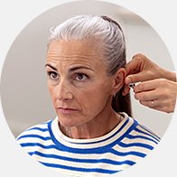 Een persoon met grijs haar draagt een gestreepte trui terwijl een audioloog een hoortoestel nauwkeurig achter het oor bevestigt in een professionele hoorzorgomgeving, mogelijk van Audika.