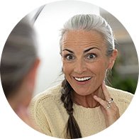 Een oudere vrouw met een grijze vlecht draagt een crèmekleurige trui en raakt haar nek aan. Ze zit in gesprek met een andere persoon in een lichte, natuurlijke omgeving.