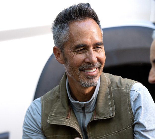 Een man in een beige bodywarmer en grijze trui praat met iemand in een buitenomgeving vlak bij een voertuig. Geen hoorapparaten of zichtbare tekst aanwezig.