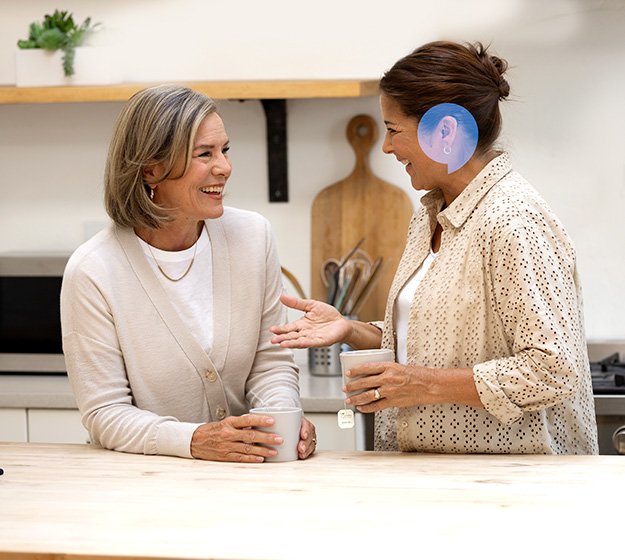 Twee vrouwen praten in een keuken terwijl ze elk een mok vasthouden; één vrouw draagt een blauw hoortoestel. Op de achtergrond staan planken met keukengerei.